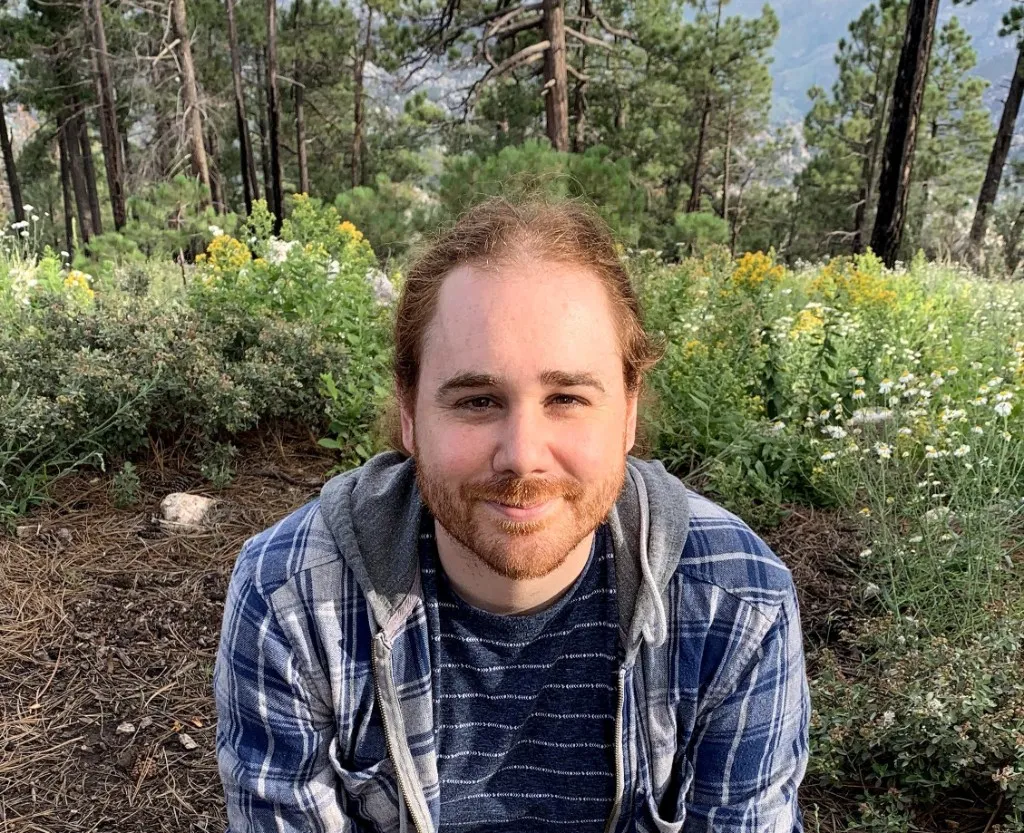 Picture of a bearded, red-haired man outdoors in a wooded setting with wildflowers, wearing a blue plaid shirt.